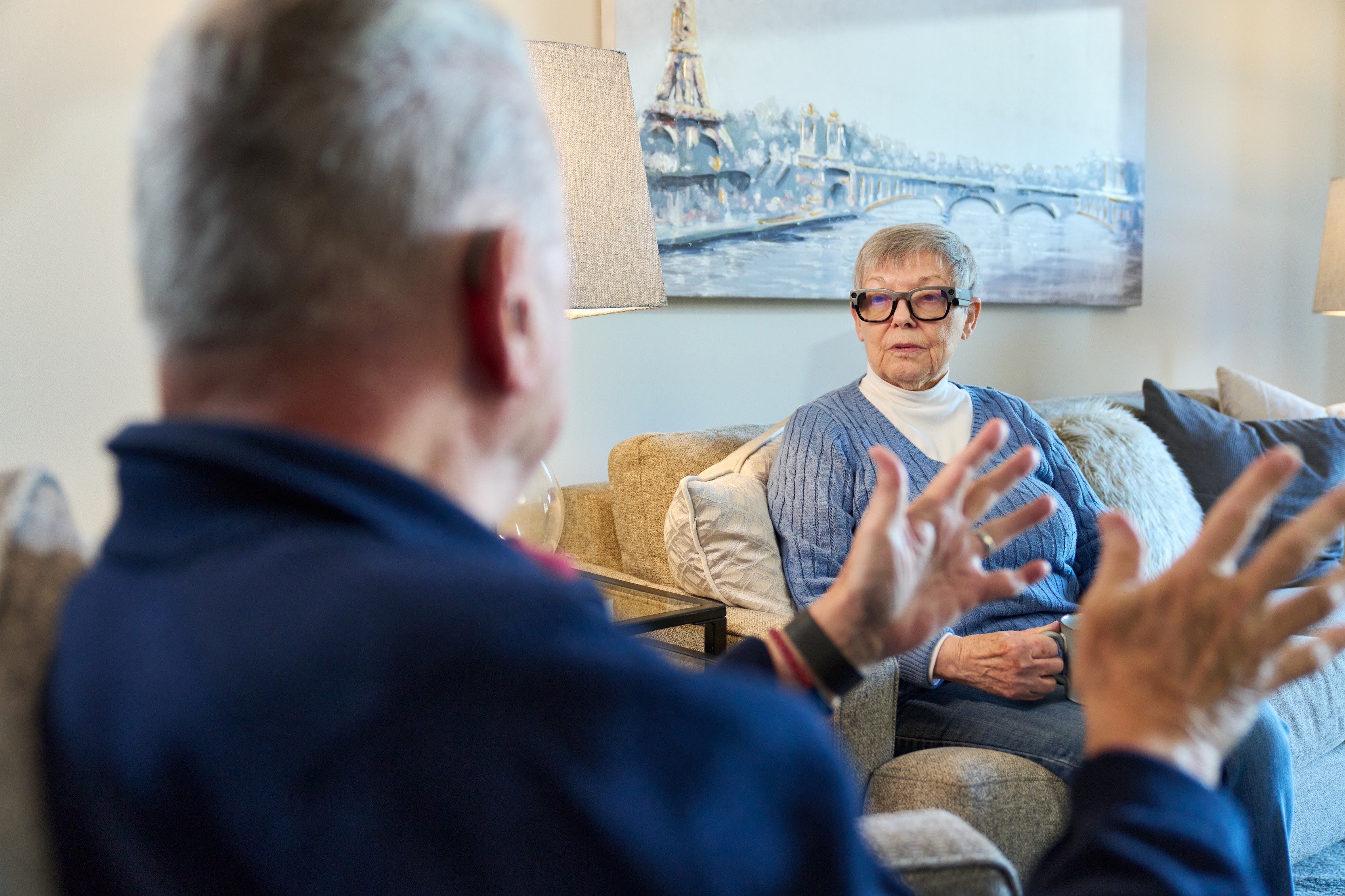 Two older adults seating facing one another. A gray-haired woman wearing glasses sits looking at a man, whose back is to the camera. He is gesturing with his hands as he talks.