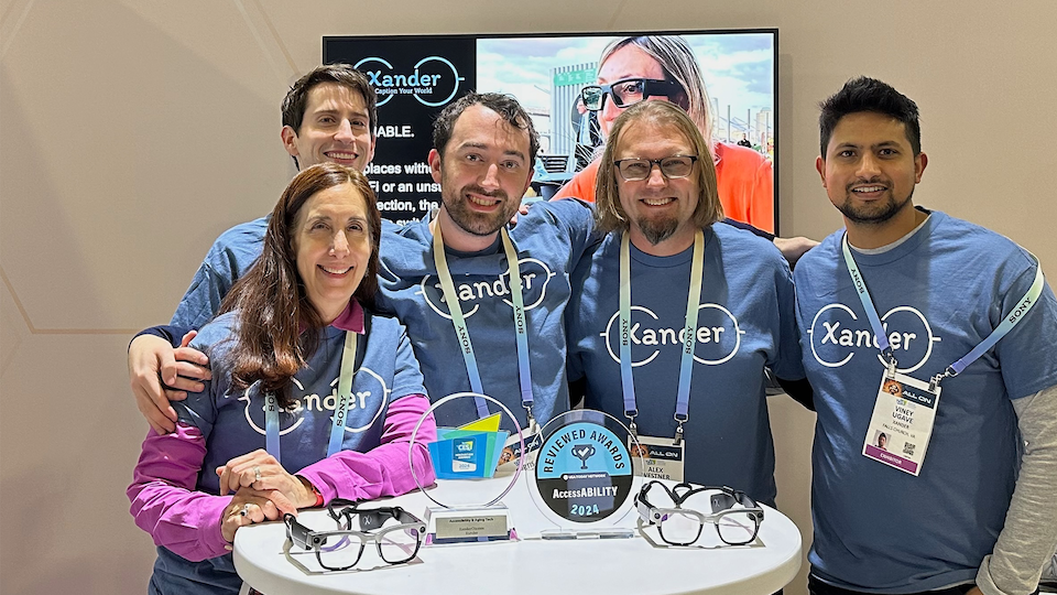 Team photo showing a woman and four men wearing blue T-shirts with company logo, Xander, standing behind a tall table that displays awards and smart glasses in front of them, at CES 2024 